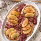 A an overhead view of boneless pork chops with apple slices and caramelized red onions arranged on top - a printed tea towel and serving tongs in the background.