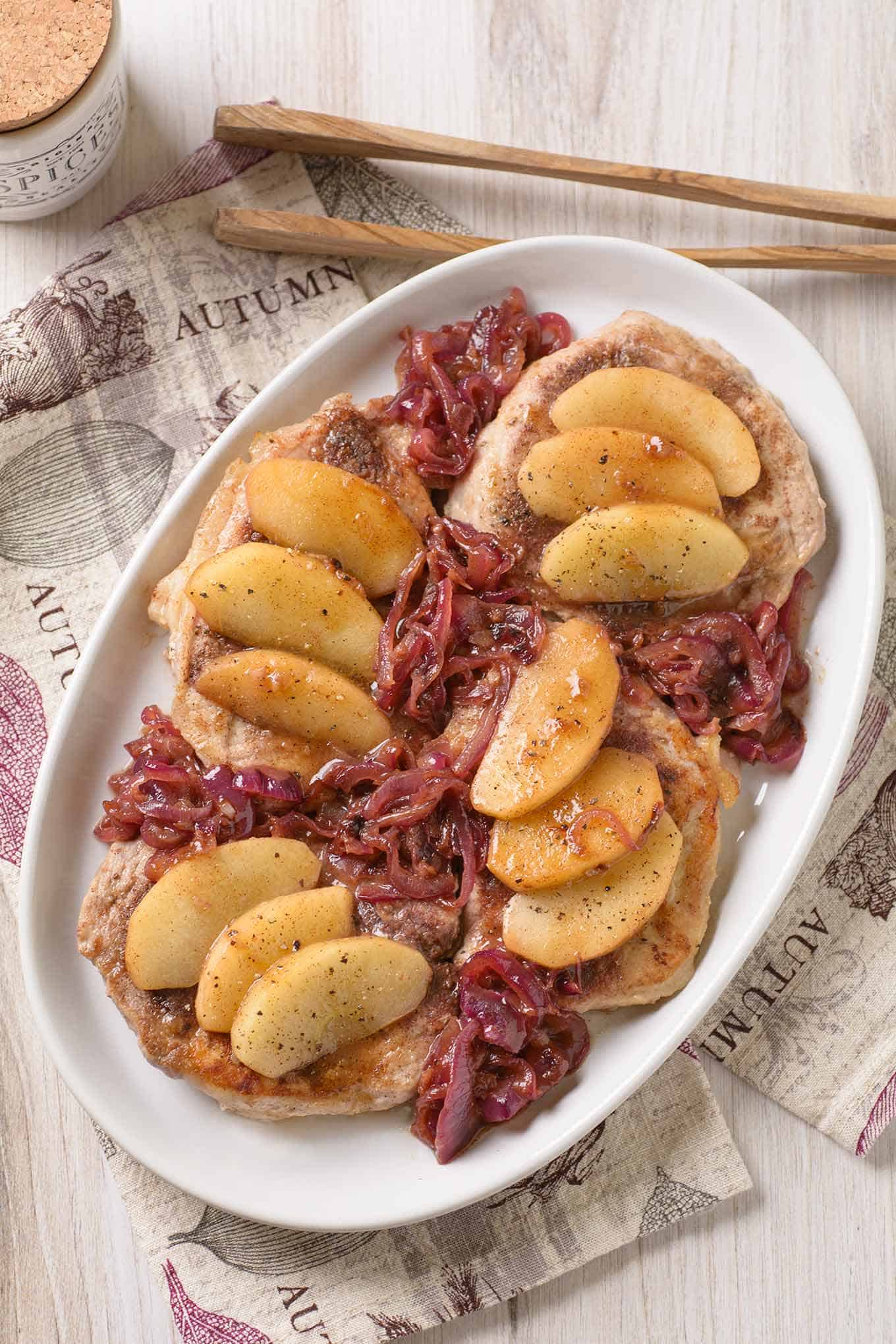 A platter with four pork chops topped with apple slices, sautéed red onion, and cider pan sauce, serving tongs on the side.