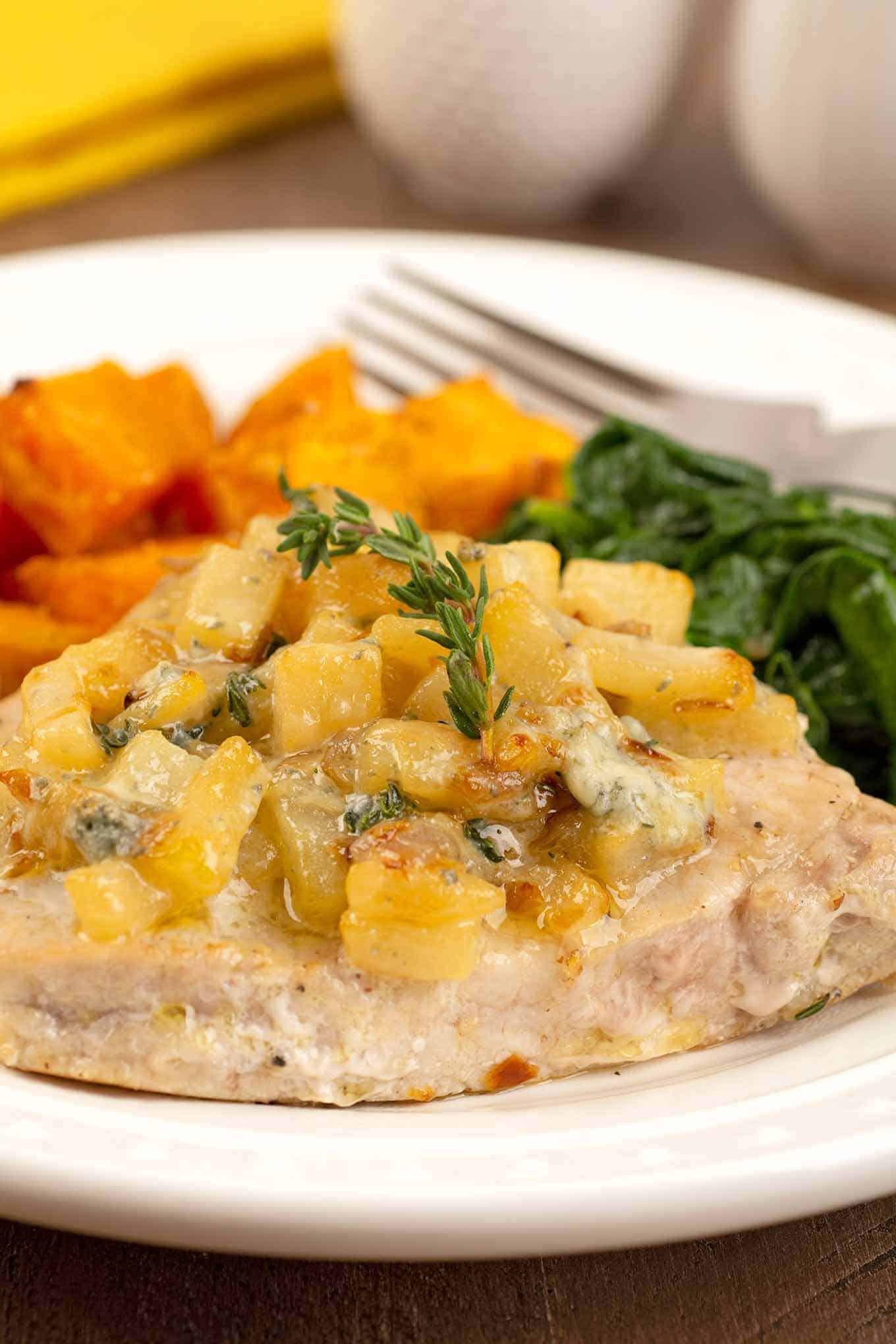 A plate with a boneless pork chop, pan-seared and topped with sautéed chopped pears, blue cheese, and thyme with side dishes of sweet potatoes and spinach.