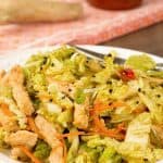 A spicy salad of napa cabbage, shredded carrots, red bell pepper, and scallions topped with stir-fried pork, a bottle of sriracha and knob of ginger on a towel in the background.