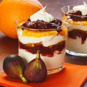 Layered parfait in a glass with mascarpone cream, sliced figs in port wine reduction, and orange segments, garnished with whipped cream, whole figs and an orange in the background.