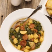 A bowl of Portuguese soup (caldo verde) made with linguica, kale, onions and potatoes. A spoon in the bowl and a plate of buttered bread in the background.