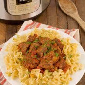 A platter of Hungarian beef goulash on a bed of wide egg noodles, with a wooden spoon and Instant Pot in the background.