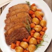 Pressure cooker pot roast served on a white platter with sliced beef, baby potatoes, and chunks of carrot, garnished with sprigs of thyme and rosemary on a rustic wooden table.