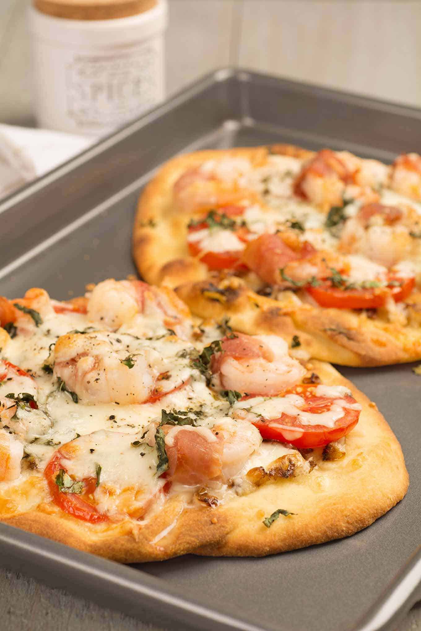 A baking sheet fresh from the oven with two naan flatbreads topped with prosciutto-wrapped jumbo shrimp, fresh tomatoes, basil, and mozzarella