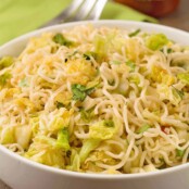 Ramen noodles tossed with sautéed napa cabbage, scallions, cilantro, and a sriracha garlic butter in a serving bowl, serving fork and bottle of sriracha in the background.