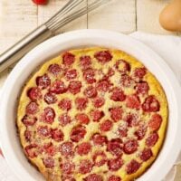 An overhead view of freshly baked raspberry clafoutis dusted with powdered sugar in a white pie plate, surrounded by a whisk, eggs, and fresh raspberries.