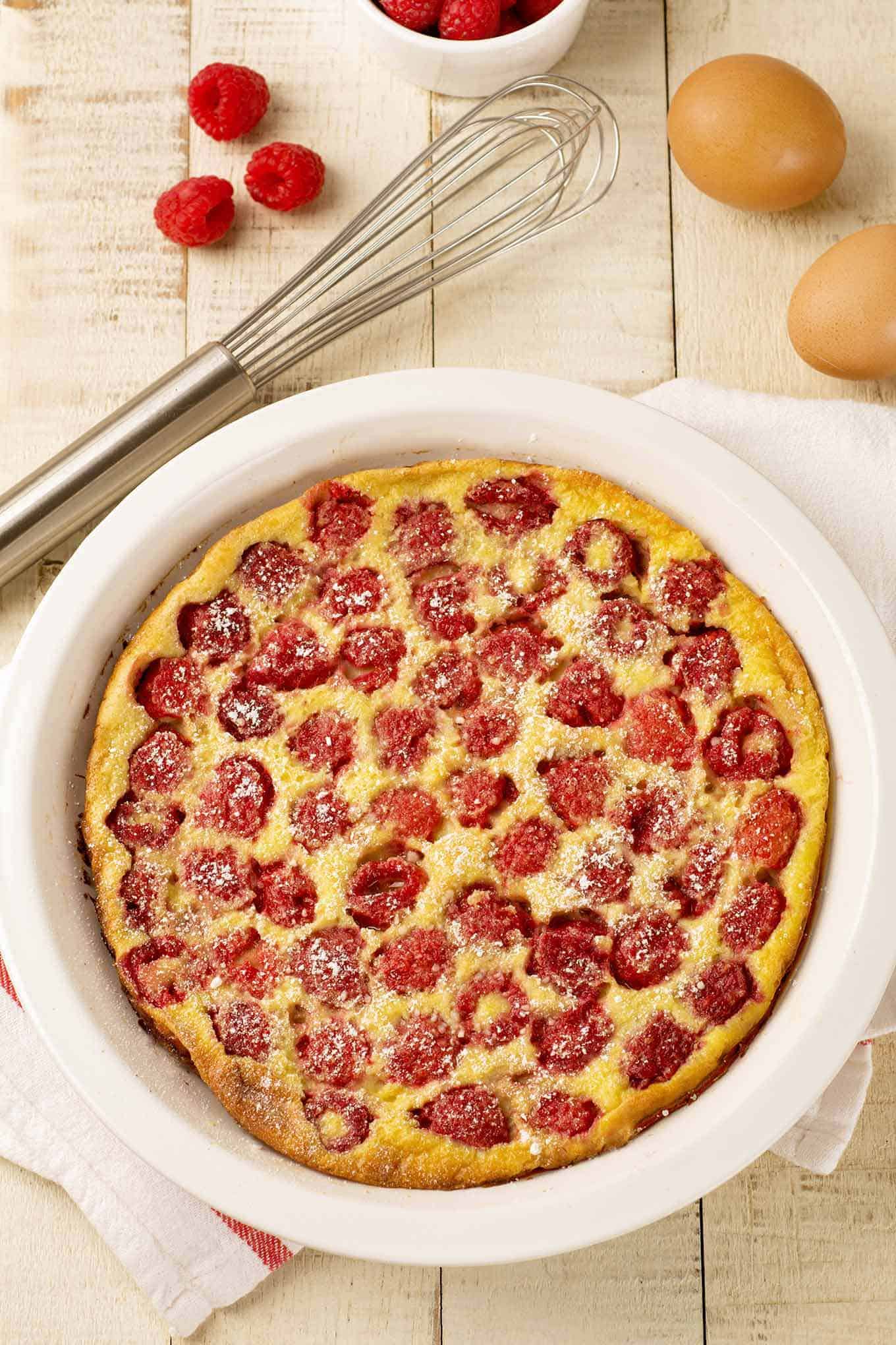 Overhead view of raspberry clafoutis with whole eggs, fresh fruit and a whisk in the background.