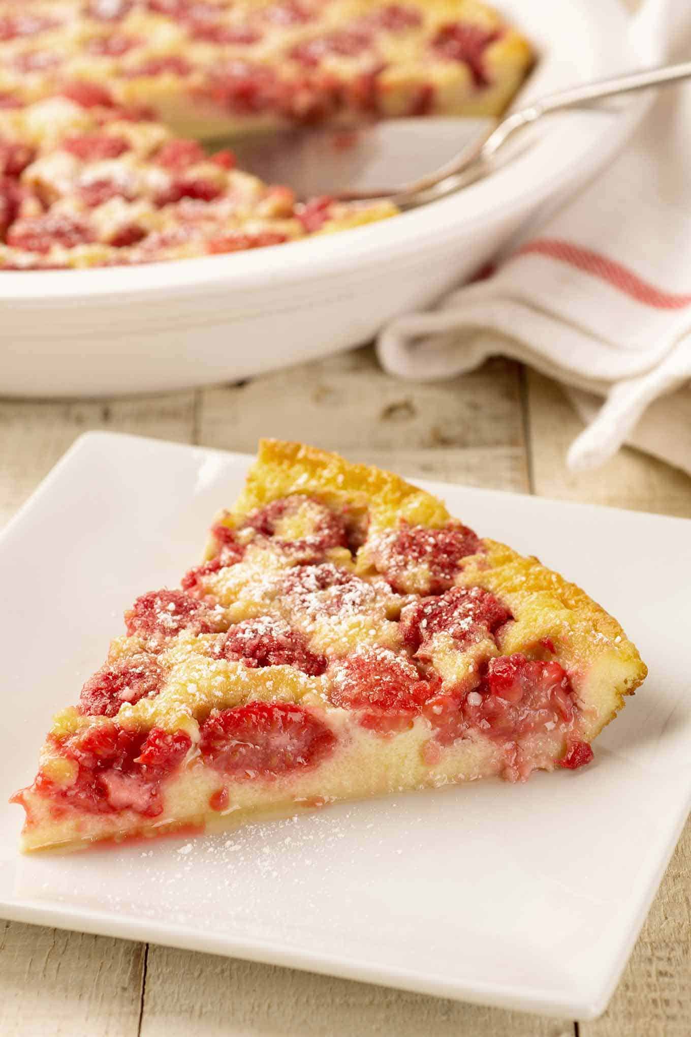 Single serving of raspberry clafoutis on a square white plate with baking dish in the background.