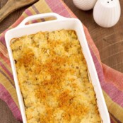 A white, oblong casserole dish of crumb-topped, riced cauliflower gratin on a tea towel with white salt and pepper shakers in the background.