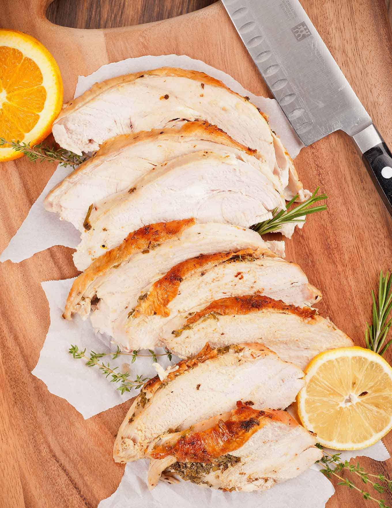 Sliced turkey breast with citrus herb butter on wooden cutting board with carving knife, fresh herbs, slices of lemon and orange