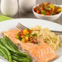 A plate with a roasted salmon fillet topped with a fresh orange, tomato, and jalapeno salsa and served with roasted green beans and quinoa, more salsa in the background.
