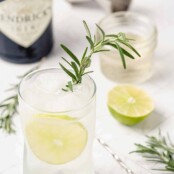 A gin and tonic cocktail made with rosemary syrup, garnished with a sprig of fresh rosemary, the gin bottle and a jar of the syrup in the background.
