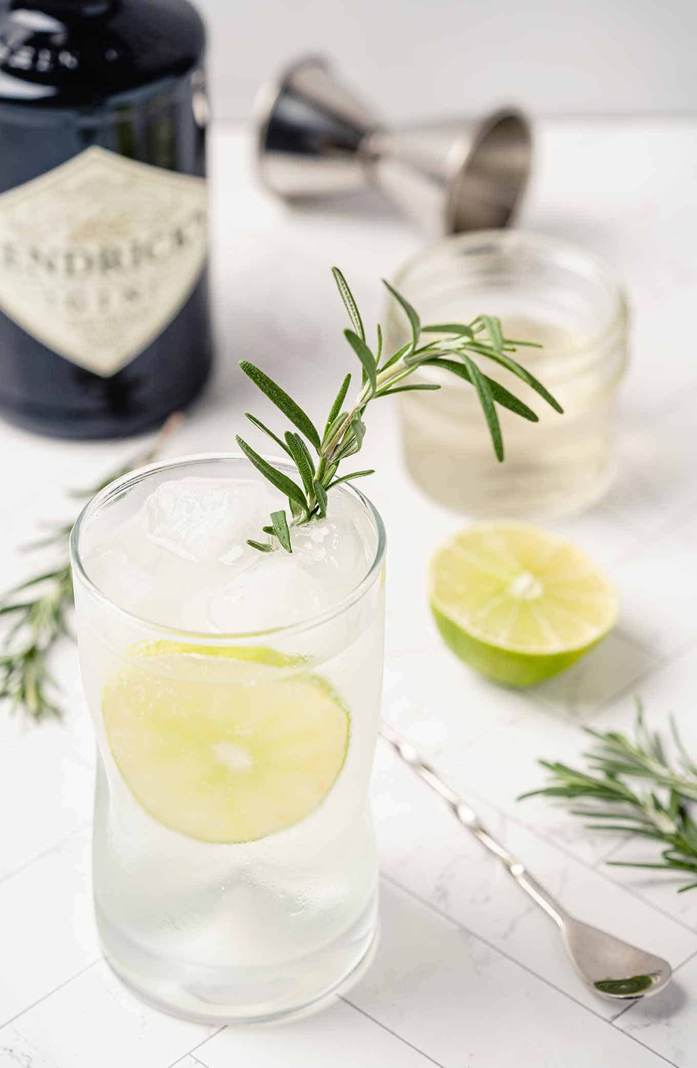 Rosemary gin and tonic garnished with lime and fresh rosemary, extra ingredients in the background.