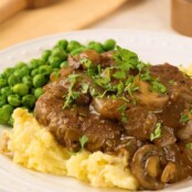 Salisbury steak topped with mushroom and onion gravy, served with mashed potatoes and peas on a white plate.