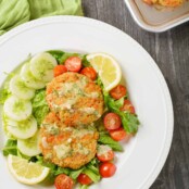 A composed green salad with lettuce, cherry tomatoes, and cucumber slices topped with lemon slices, fresh dill, two salmon cakes, and dill dressing.