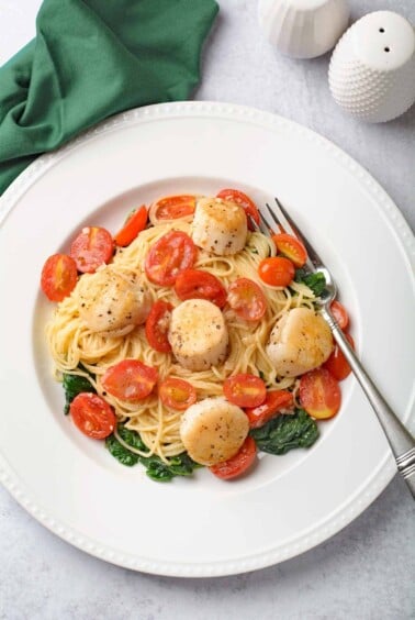 Scallops and pasta with white wine butter sauce, angel hair, tomatoes, and saut&eacute;ed spinach on a white plate.
