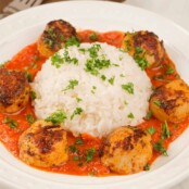 A plate of paprika-crusted, pan-seared sea scallops in roasted red pepper sauce arranged around a serving of white rice, all garnished with chopped parsley.