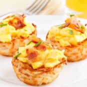A serving plate with three hash brown potato cups filled with scrambled eggs topped with bacon and garnished with chives.