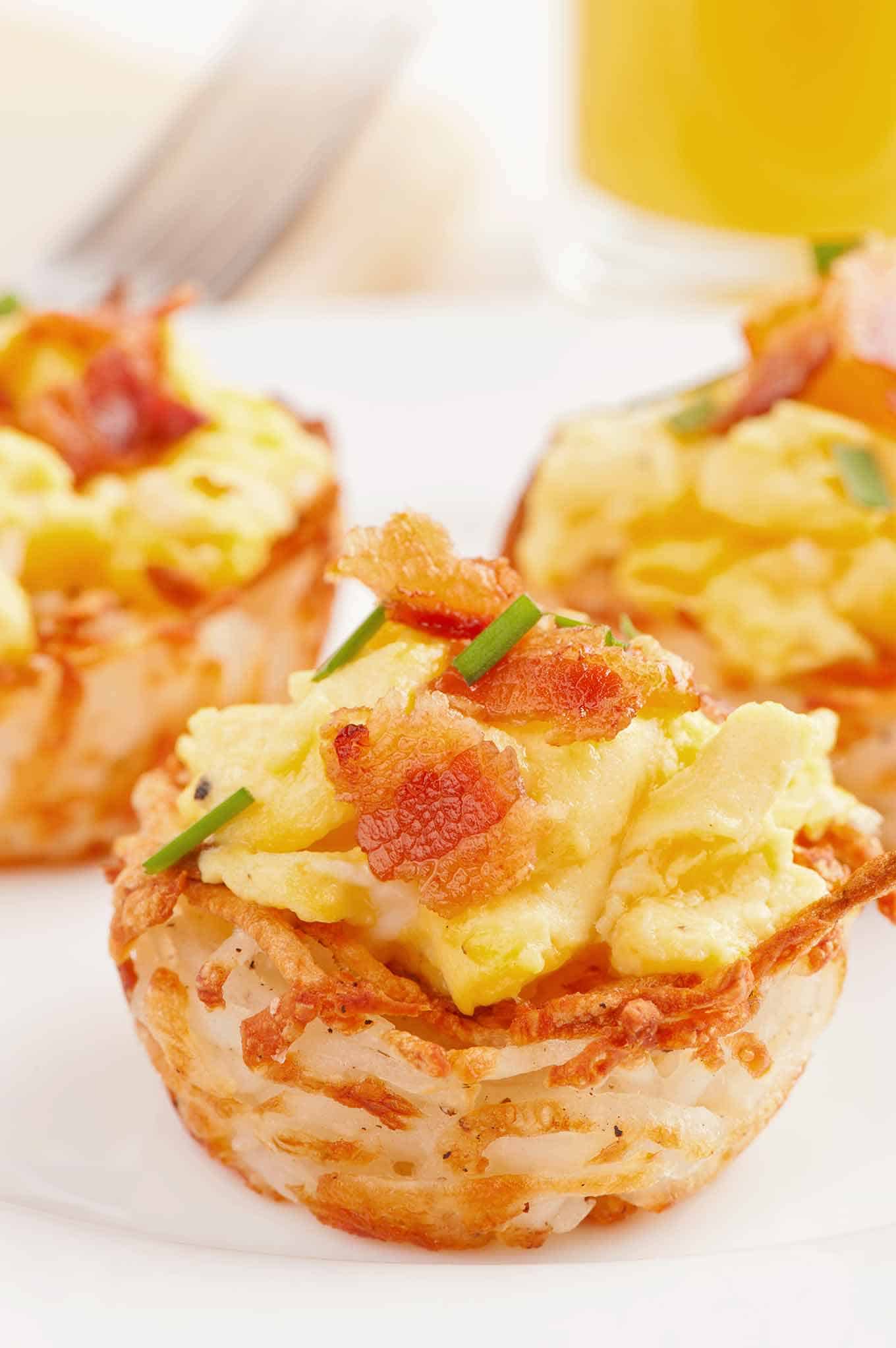 Scrambled eggs inside hash browned potato cup topped with bacon and garnished with chives.