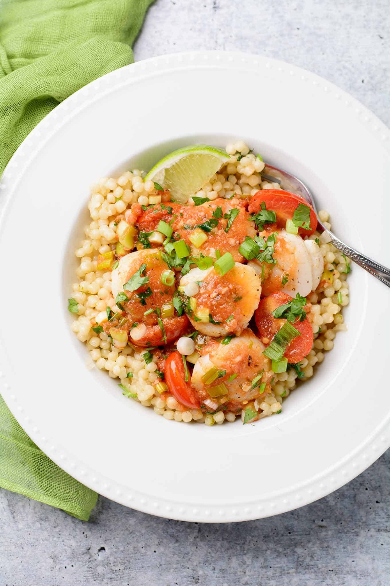 A serving of pearl couscous topped with seared scallops, grape tomatoes, and scallions in tomato-lime butter.