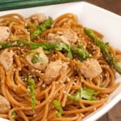 Bowl of sesame noodles mixed with chunks of chicken and asparagus spears, garnished with sesame seeds and fresh herbs, chopsticks resting on the edge of the bowl.