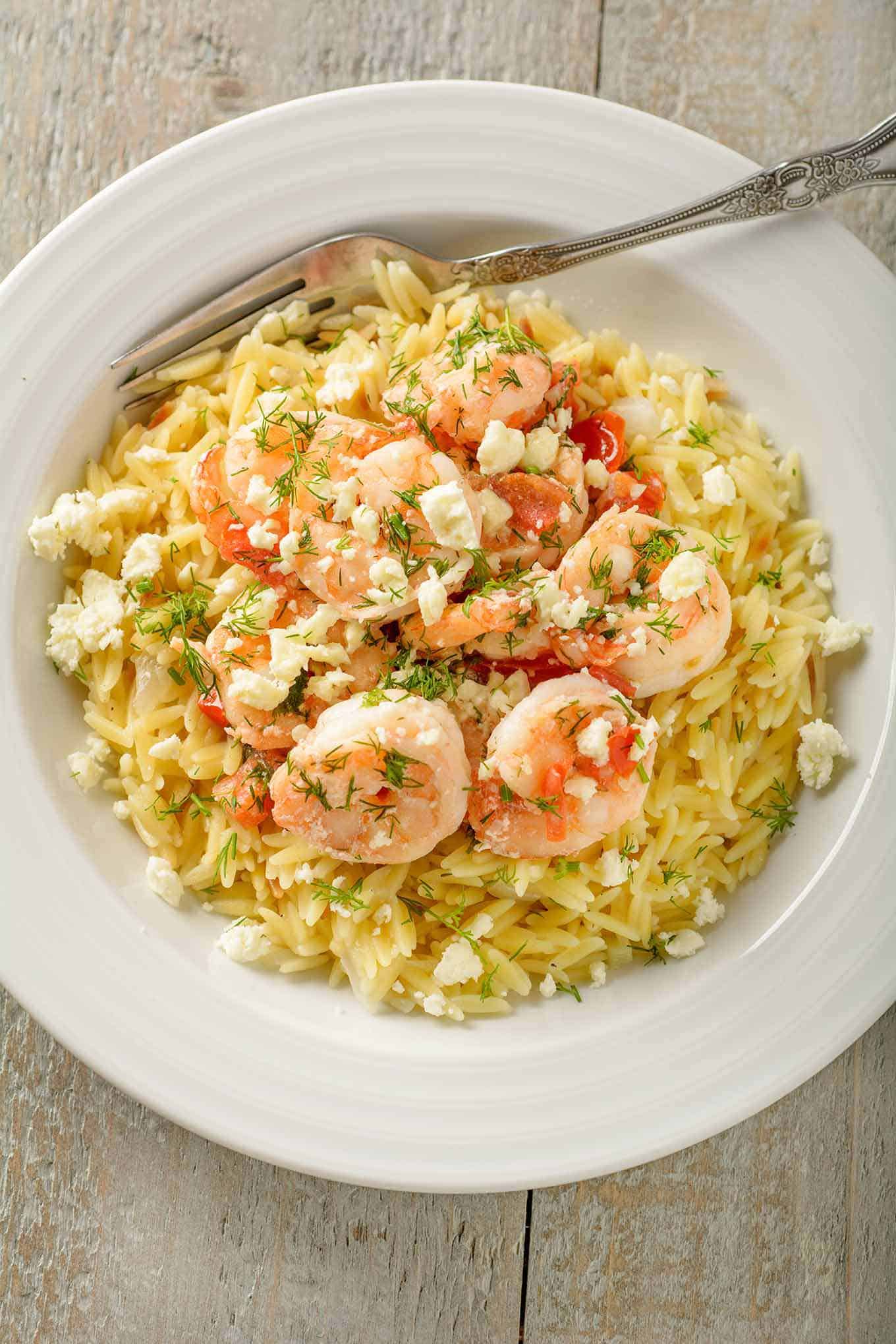 An overhead view of a bowl of Greek style sautéed shrimp with dill, lemon, diced tomatoes, and feta cheese, served over orzo.