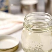 A jar of prepared simple syrup with the lid beside it, bag of sugar, measuring cup, and saucepan in the background