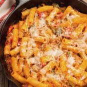 Skillet baked ziti with sausage and bell peppers, topped with melted cheese and served in a cast iron pan on a rustic wooden table.