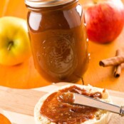 An English muffin on a cutting board being spread with homemade, slow cooker apple butter, a Mason jar filled with apple butter in the background, alongside whole apples and cinnamon sticks.