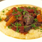 Italian pot roast served over creamy polenta, topped with tender carrots, stewed tomatoes, and chopped parsley on a white plate.