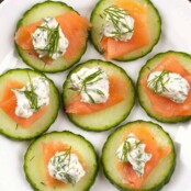 A serving plate filled with cucumber rounds topped with smoked salmon, cream cheese, and fresh dill.
