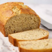 A partially sliced loaf of sour cream banana bread on a marble board with a bread knife in the background on the right.