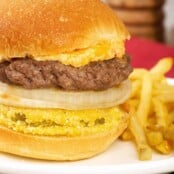 A close view of a grilled burger stacked on a bun with a slices of fried green tomato and sweet onion, and a topping of pimento cheese - fries on the side.