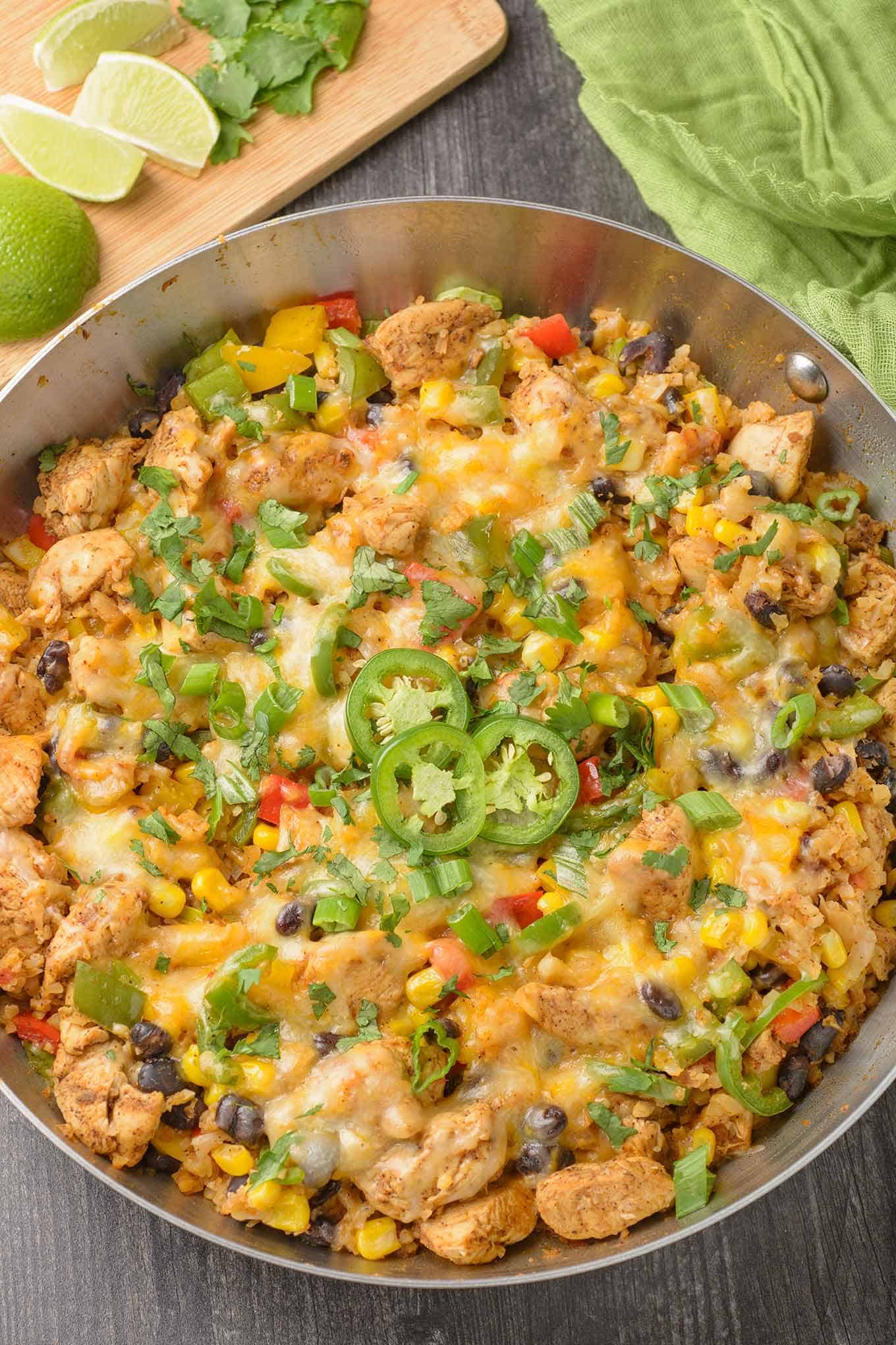 A frying pan filled with Southwest-style chicken and cauliflower rice with bell peppers, corn, scallions, beans, garnished with sliced jalapenos and cilantro.