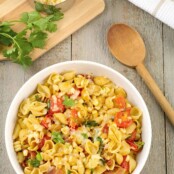 A serving bowl of pasta shells with bacon, corn, tomatoes, chipotle, cheese, and Southwest seasonings, with a cutting board, cilantro, and spoon to the side.