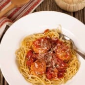 A plate of spaghetti topped with meatballs in marinara sauce, garnished with grated Parmesan cheese, on a wooden table with a napkin, wooden spoon, and a bottle of Chianti.