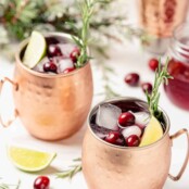 Two copper mugs filled with Moscow Mule cocktail, along with ice cubes, fresh cranberries, lime wedges and sprigs of fresh rosemary.