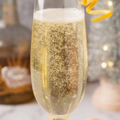 A sparkling wine cocktail in a champagne flute garnished with an orange twist, orange slices.