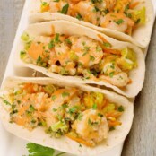 An overhead view of soft flour tortillas filled with crisp napa cabbage slaw, jumbo shrimp stir-fried with garlic and ginger, and a garnish of chopped cilantro.