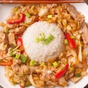 A white plate of spicy stir-fried pork with cabbage, red bell peppers, and scallions served around a mound of steamed white rice, with a pair of chopsticks resting on top.