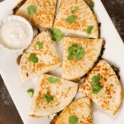 An overhead view of a platter of spinach, mushroom, and cheese quesadillas garnished with cilantro, a dipping bowl of sour cream in the corner.