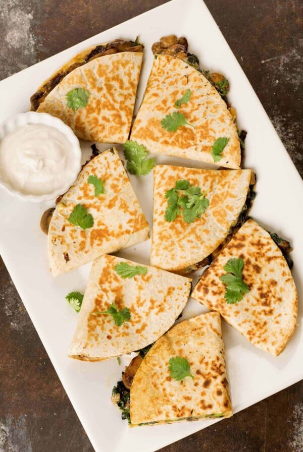 An overhead view of a platter of spinach, mushroom, and cheese quesadillas garnished with cilantro, a dipping bowl of sour cream in the corner.