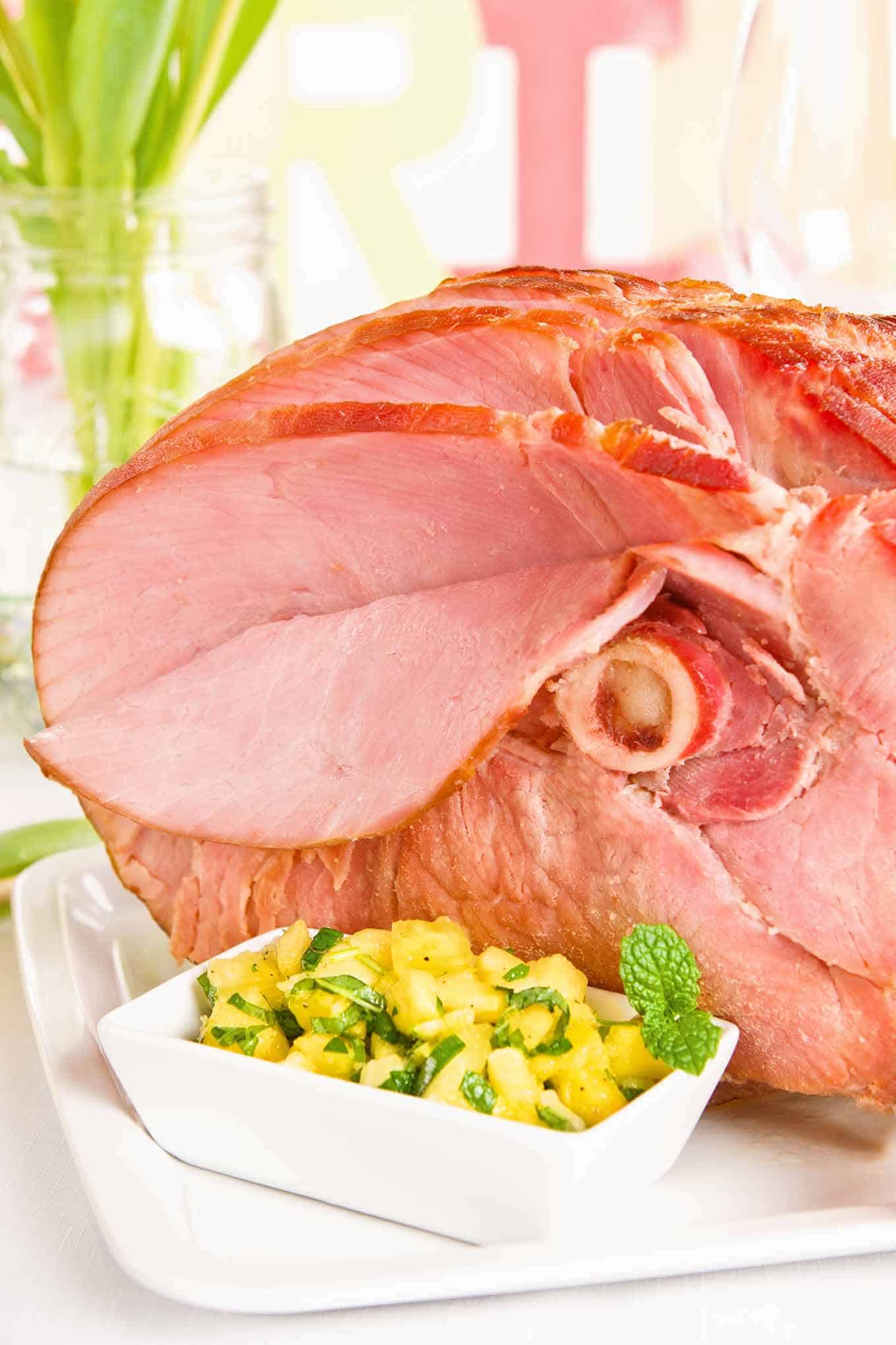 A spiral-sliced, glazed, baked ham glazed on a serving platter with an accompanying bowl of pineapple mint chutney, surrounded by spring table decor.