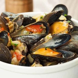 A white serving bowl filled with steamed mussels, bits of tomato and onion, and chopped parsley.