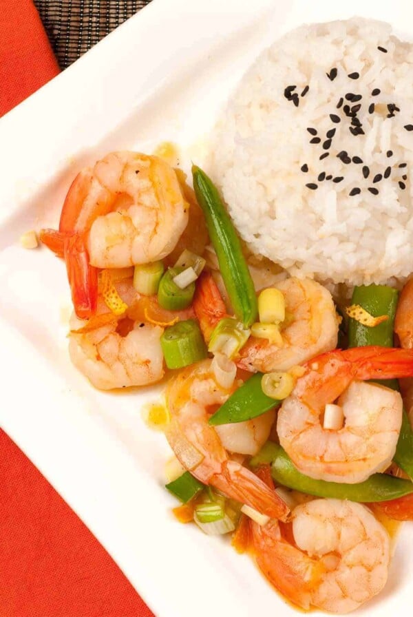 An overhead view of stir-fried shrimp with orange peel, snap peas, and scallions served with white rice topped with black sesame seeds.