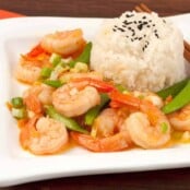 A side view of stir-fried shrimp with orange peel, snap peas, and scallions served with white rice topped with black sesame seeds.