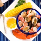 Plate containing lobster claw, ear of corn, bowl of melted butter, nutcracker, and bowl of steamed clams, mussels, sausage, and potatoes.