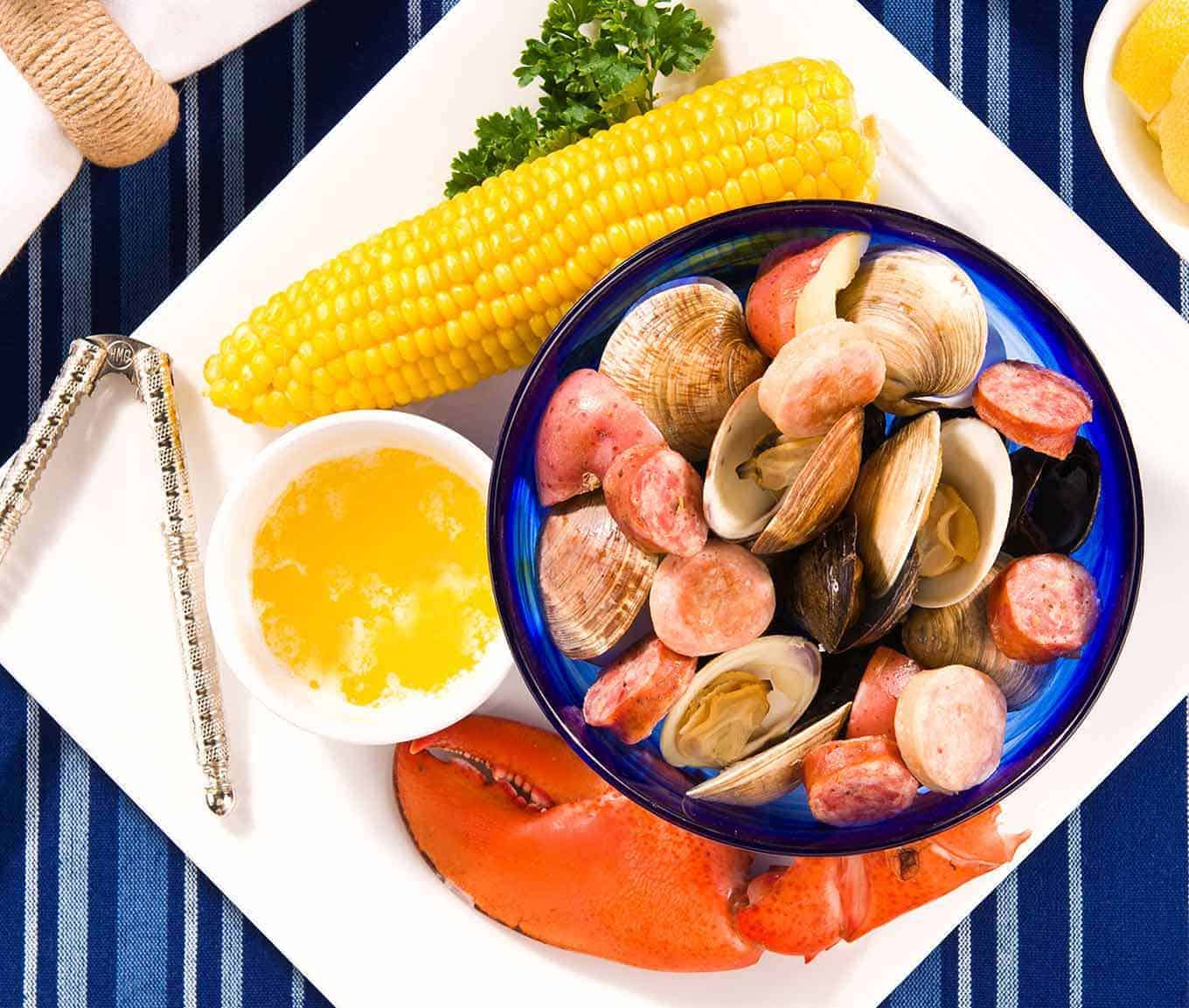 Plate containing lobster claw, ear of corn, bowl of melted butter, nutcracker, and bowl of steamed clams, mussels, sausage, and potatoes.