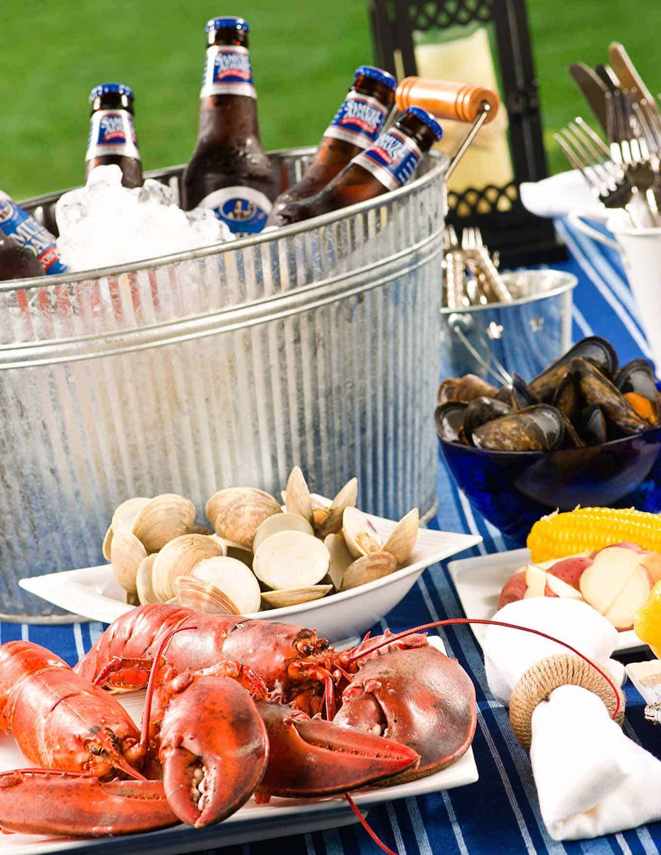 Table filled with with lobsters, clams, mussels, potatoes, corn, utensils, and an ice-filled bucket with cold bottles of beer.